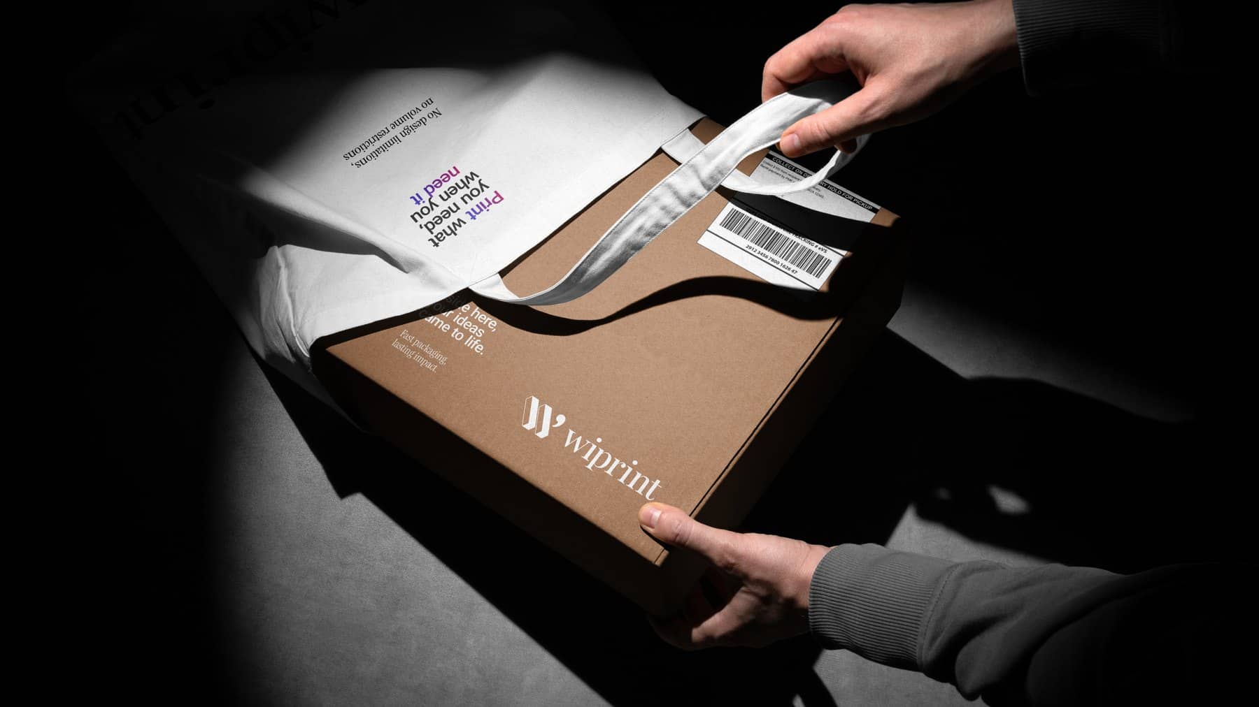 Hands closing a kraft mailer box printed with the WiPrint logo, beside a printed garment bag.